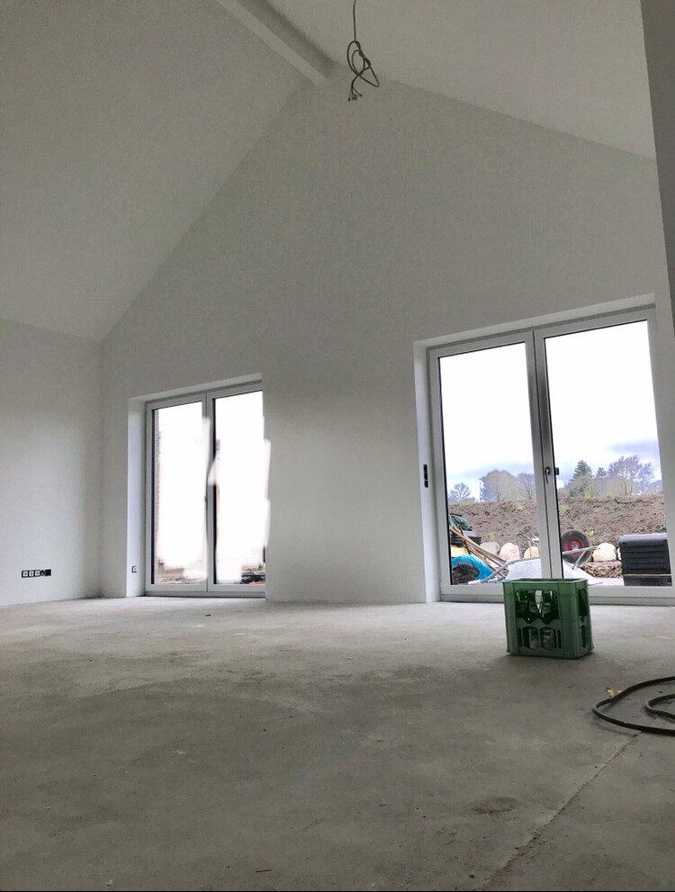 Heller, leerer Neubau-Raum mit hohen Decken, großen Glasfenstern; Baugeräte draußen.