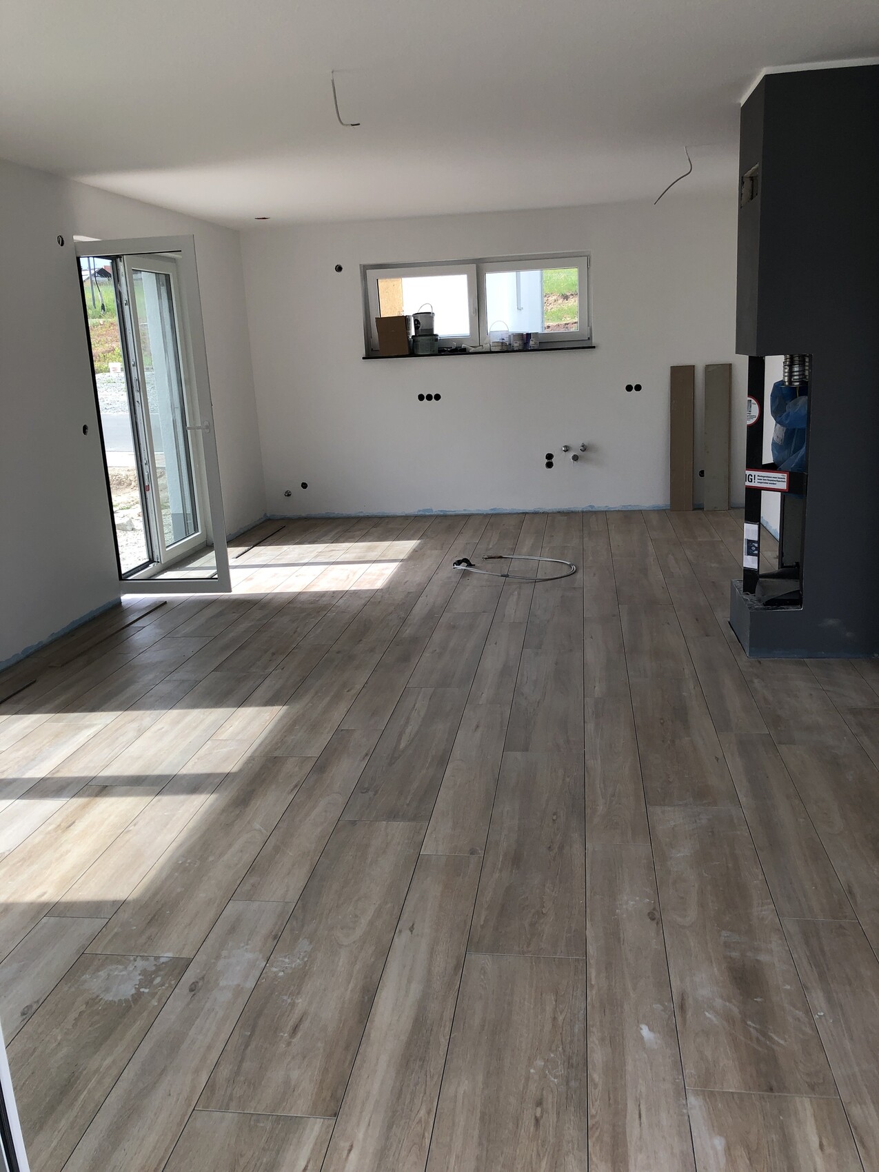 Heller unfertiger Wohnraum mit Holzoptikboden, weißen Wänden, Fensterfront und sichtbaren Kabeln.