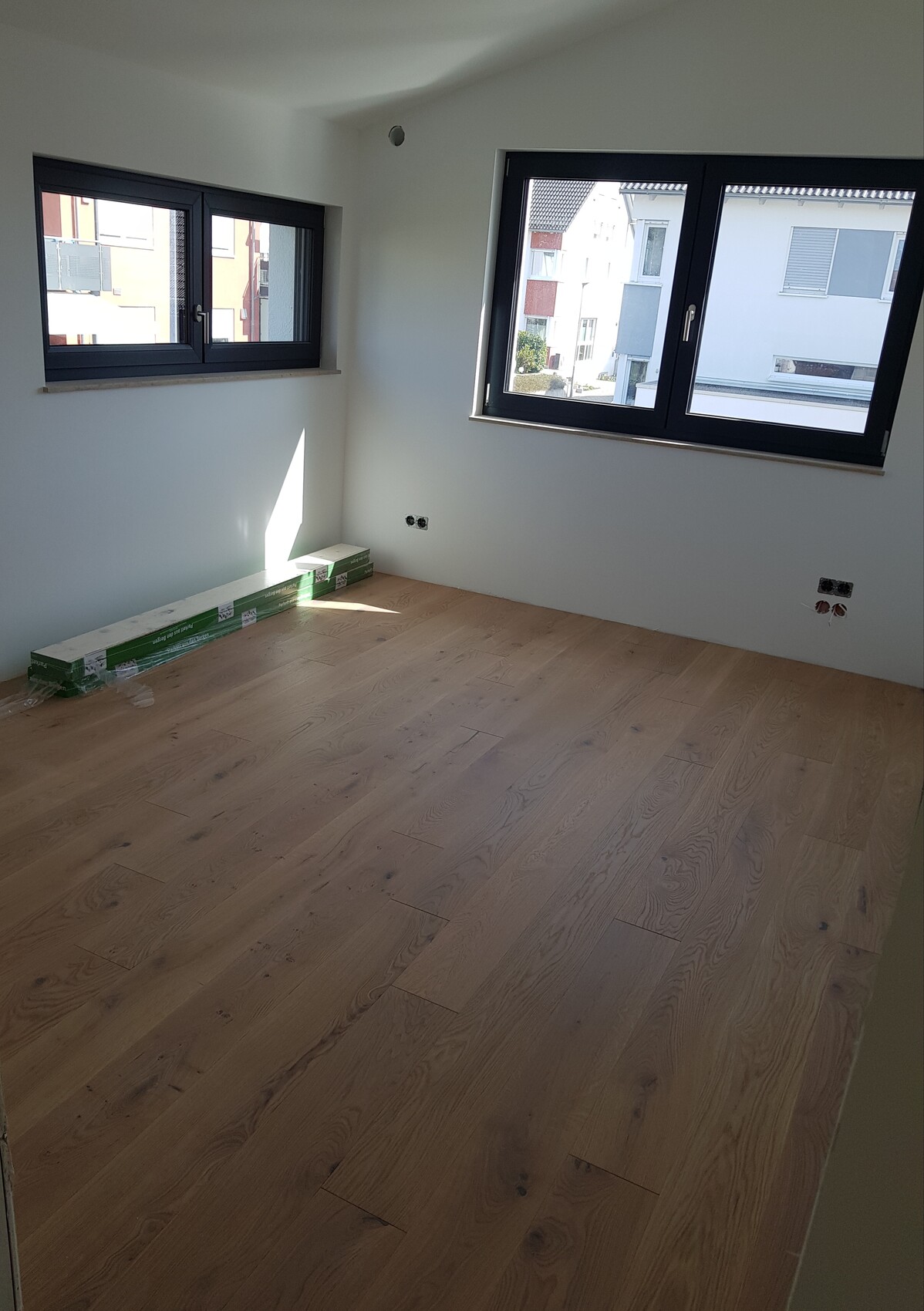 Leerer Raum mit Holzfußboden, zwei großen Fenstern und noch verpacktem Laminat am Boden.