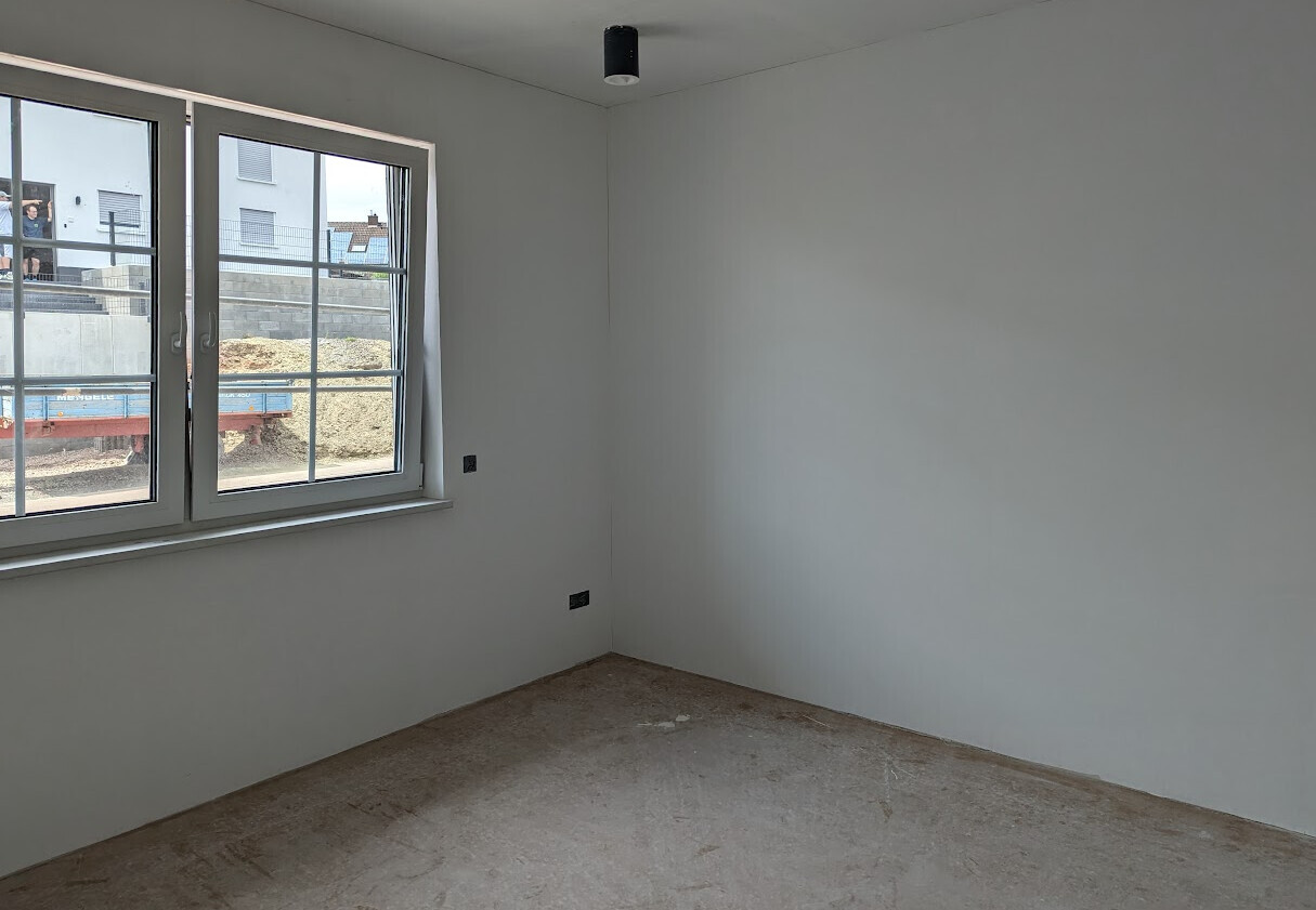 Leerer Renovierungsraum mit großem Fenster, Betonboden, weißen Wänden und Deckenlampe.
