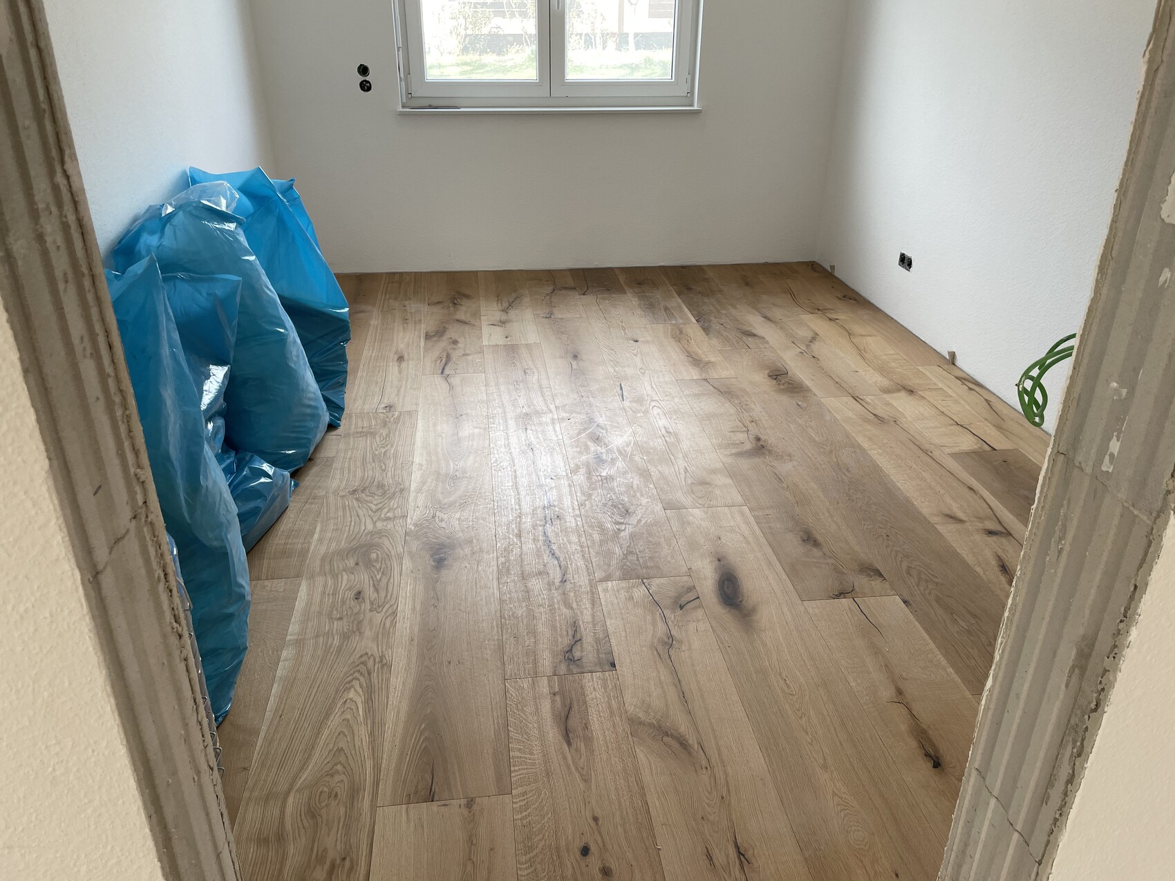 Leerer Raum mit Holzboden, blaue Müllsäcke links, Fenster an der Rückwand, weiße Wände.