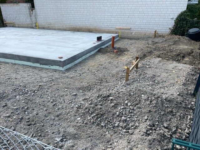 Baustelle: neue Betonfundamentplatte links, Erdboden mit Kies, Rohre und Markierungen sichtbar.