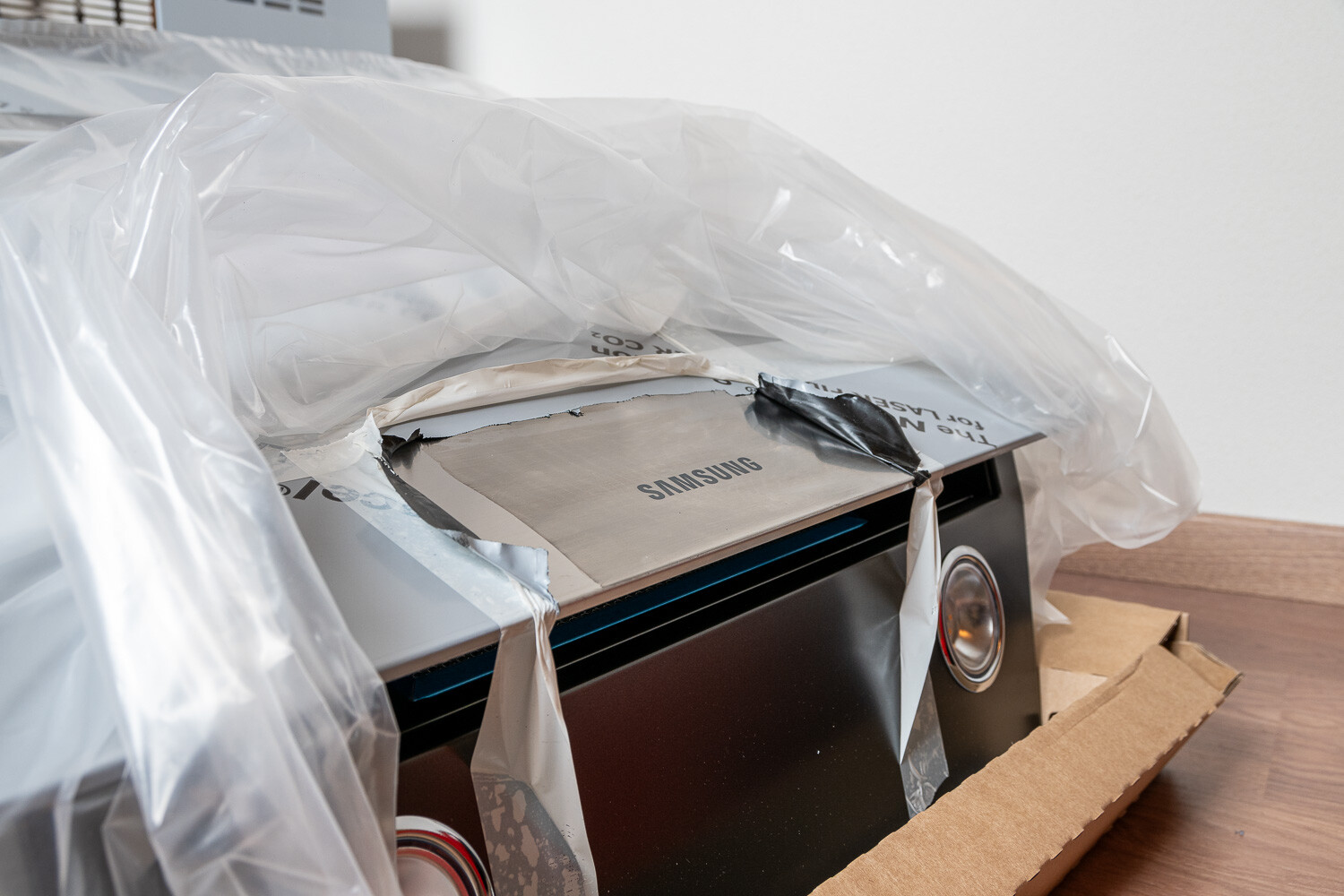 Verpacktes Samsung-Gerät mit Plastikfolie darüber, Metallfront und Rundknopf; steht auf Karton.