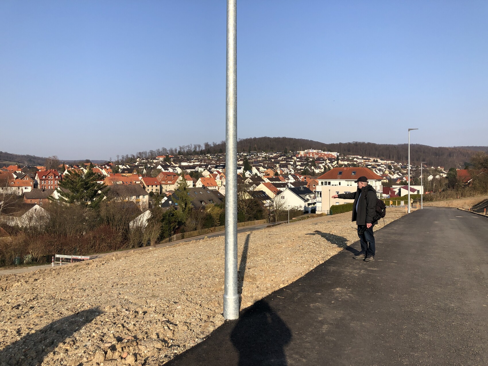 Mann mit Rucksack steht am Straßenrand und zeigt auf eine Häuserstadt am Hang unter blauem Himmel