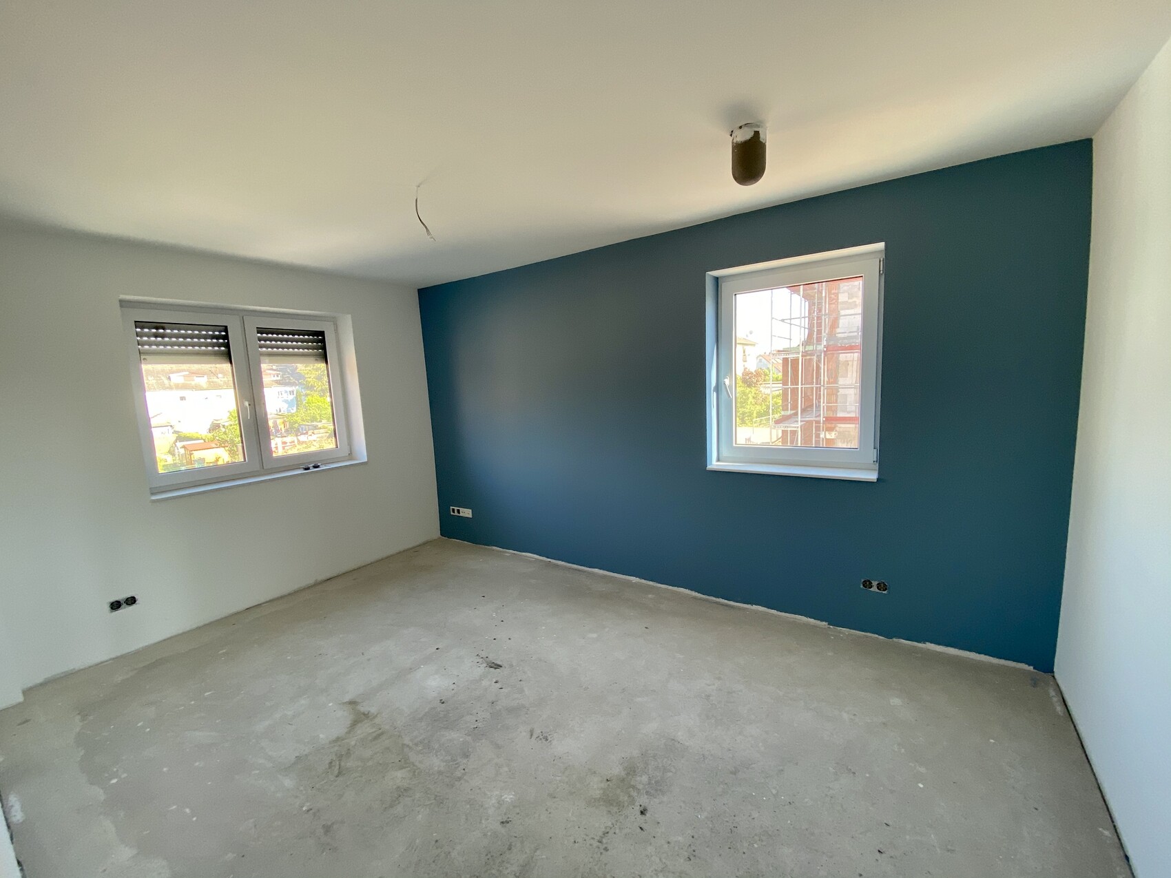 Unfertiges Zimmer mit blauer Akzentwand, zwei Fenster, rohem Betonboden.