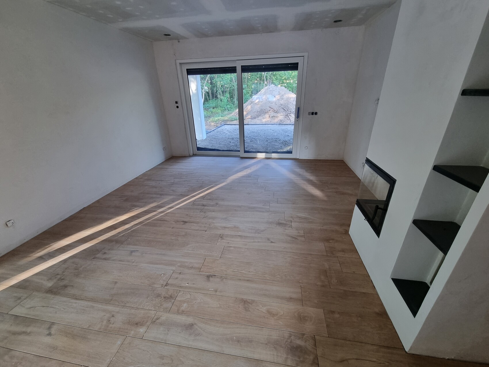 Leerer Wohnraum mit Holzboden, Kamin rechts, Glas-Schiebetür nach draußen; Baustelle sichtbar.