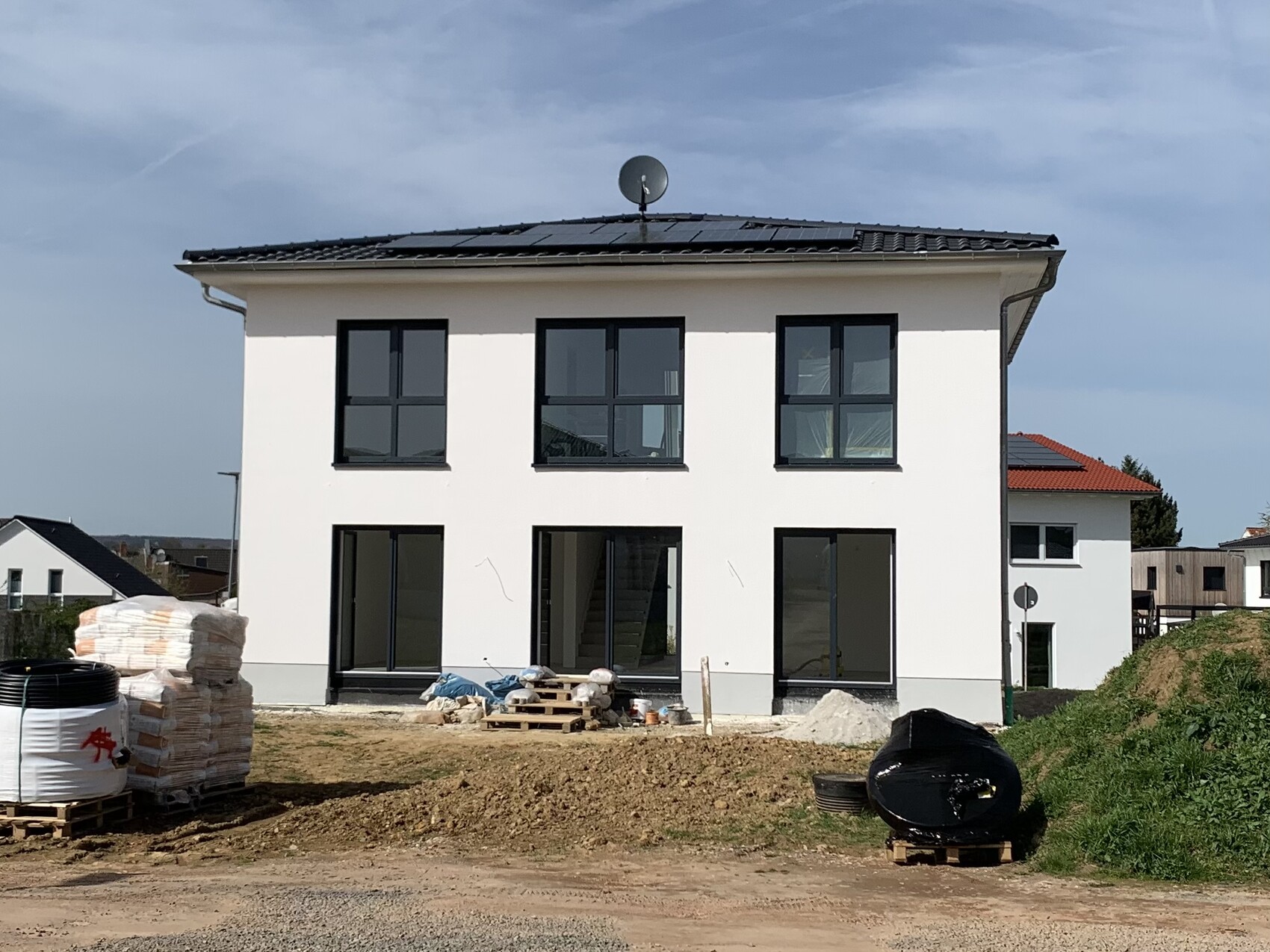 Zweistöckiges, weißes Neubauhaus mit schwarzen Fenstern; Baustellenmaterial vor dem Haus.