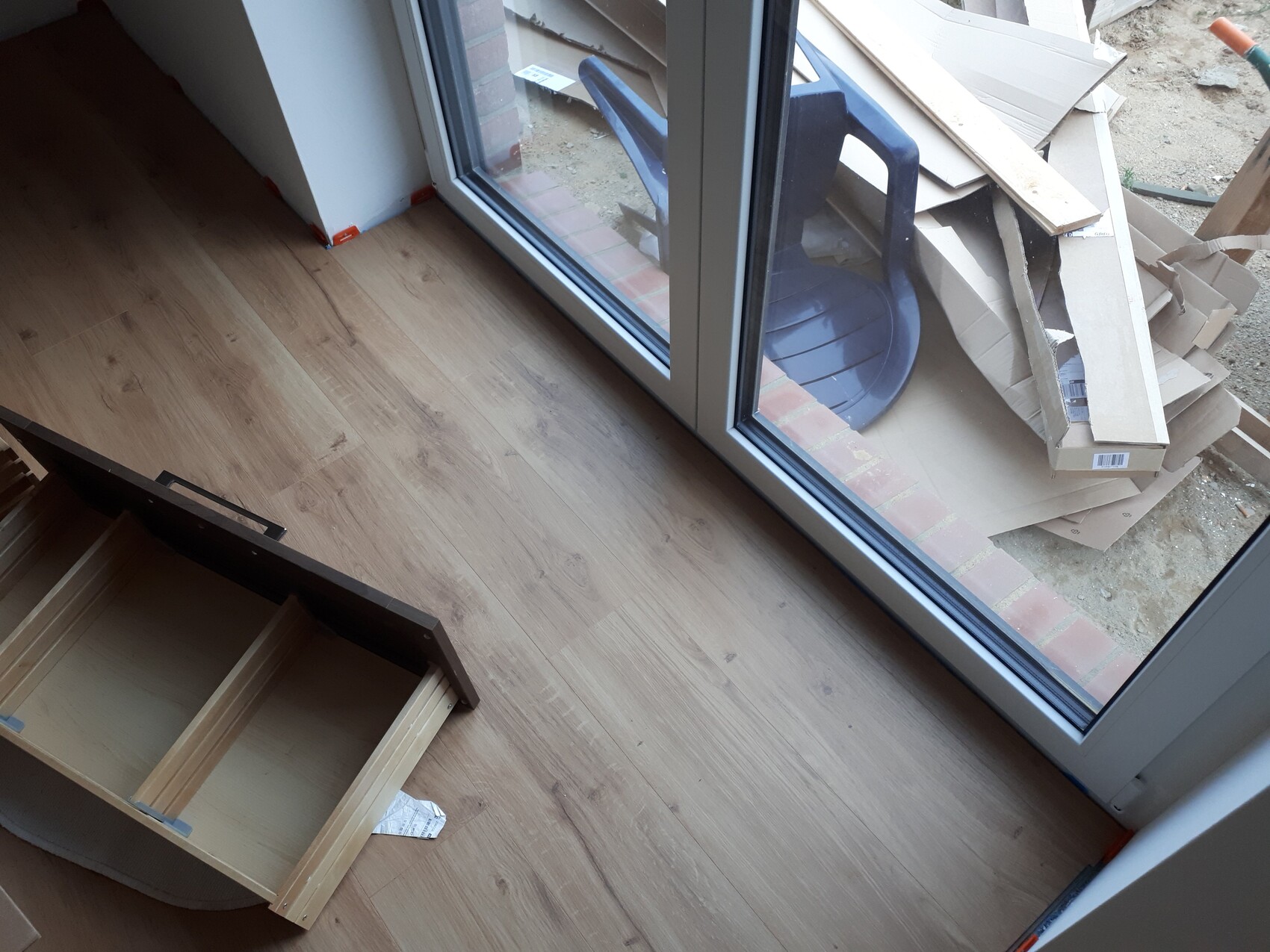 Innenraum mit neuem Holzboden; Glasschiebetür außen, Bauabfälle und eine Schublade auf dem Boden.