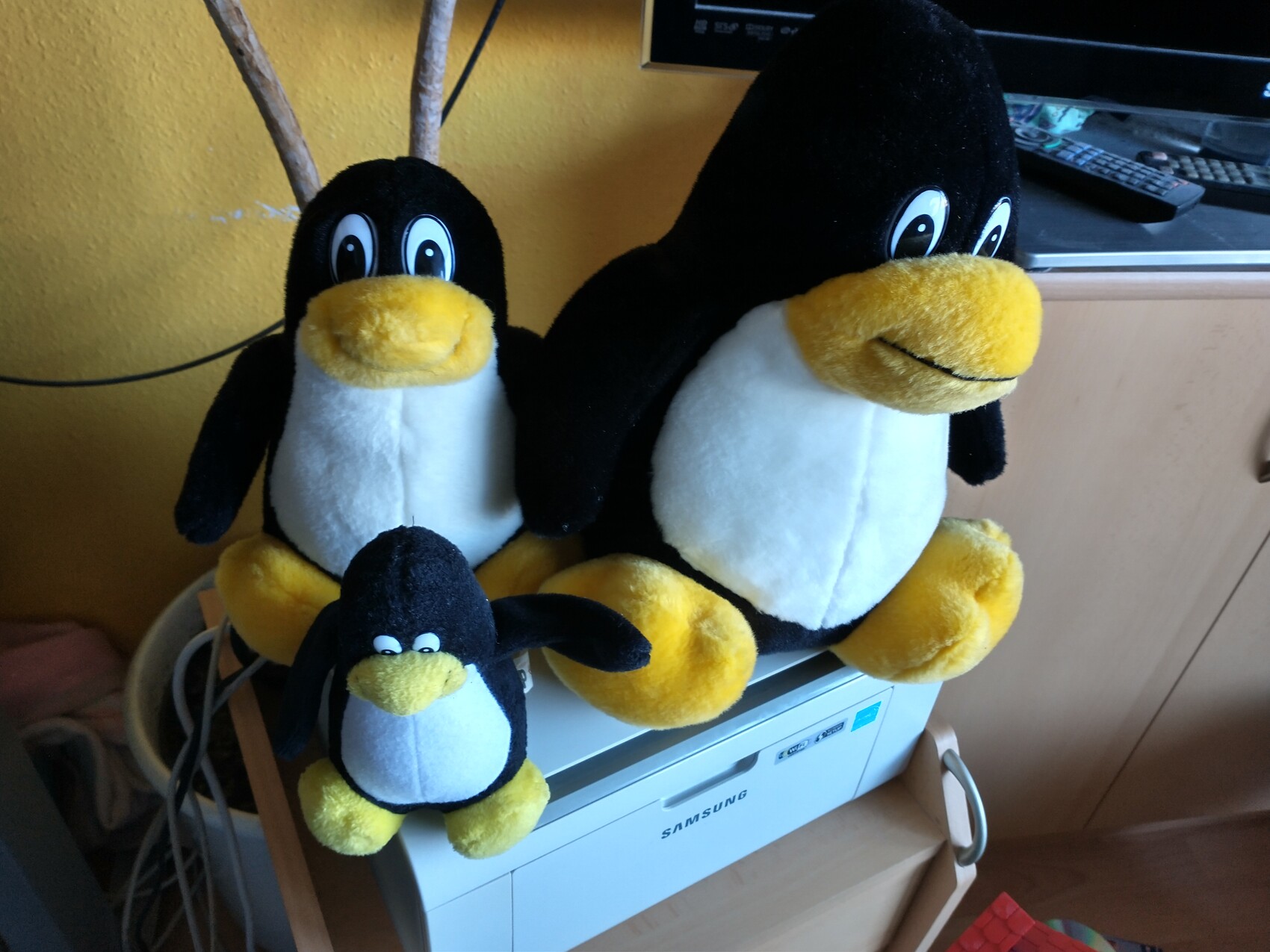 Drei Plüschpinguine sitzen auf einem Samsung-Drucker vor einer gelben Wand.