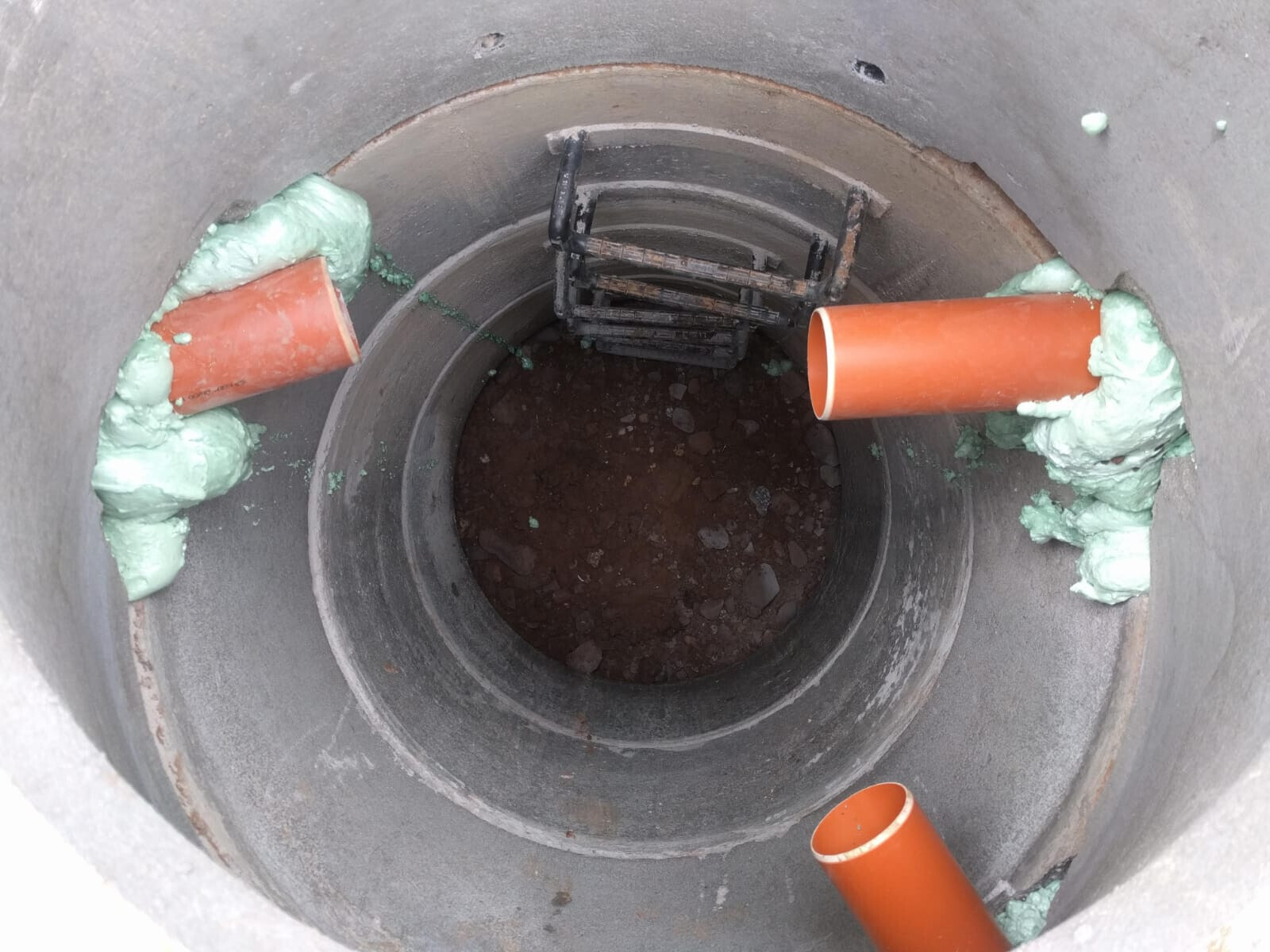 Offene Schachtöffnung mit orange PVC-Rohren, grünem Dichtstoff und Metallgitter.