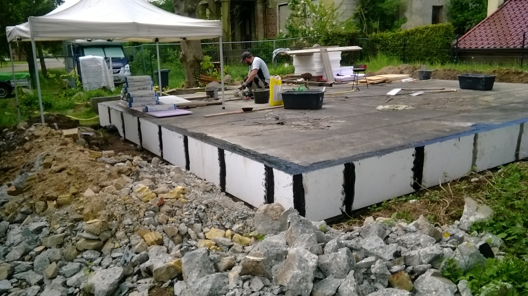 Baustelle: Fundamentplatten werden gesetzt; Arbeiter arbeitet unter Zelt mit Baumaterialien.