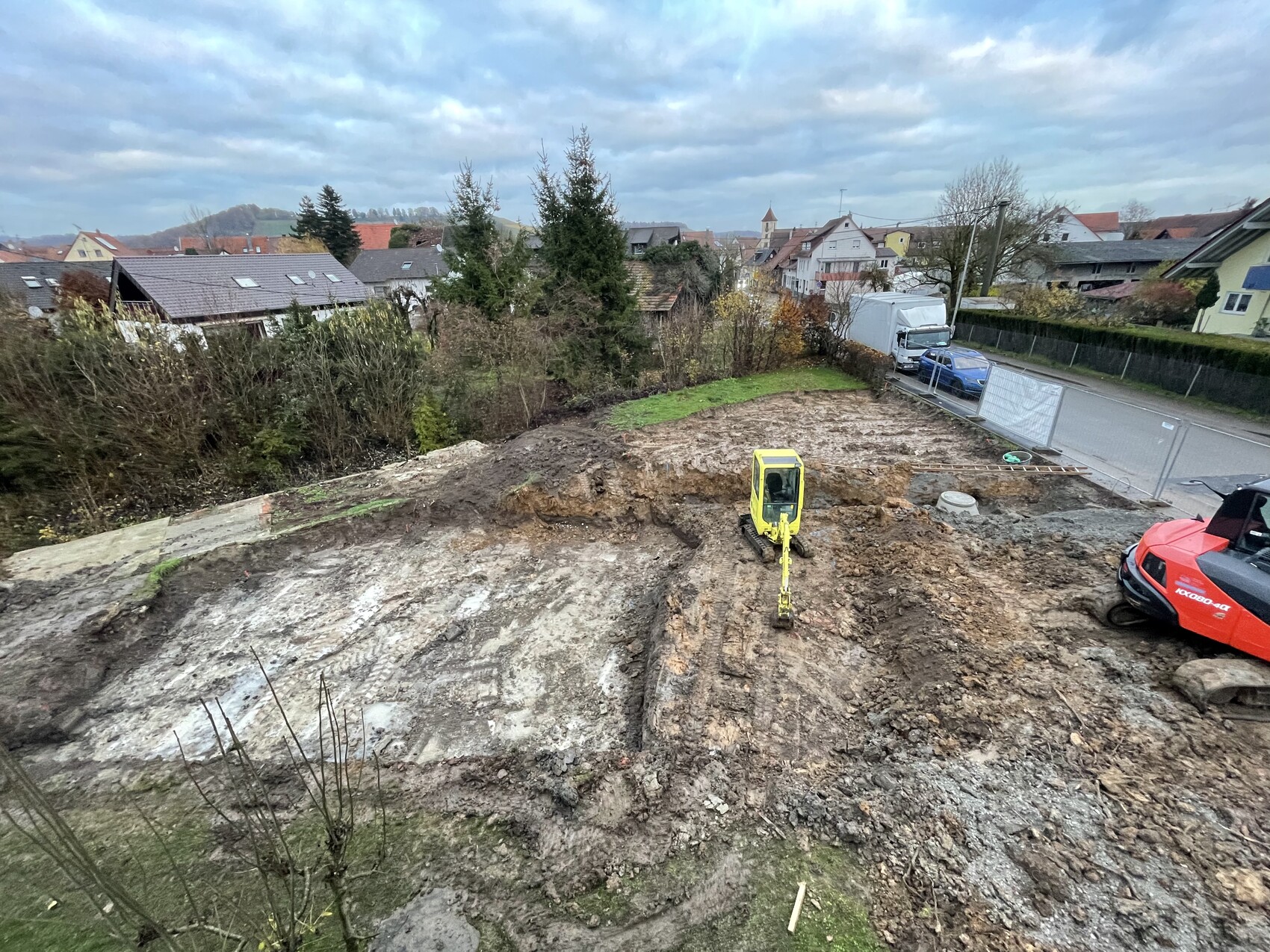 Baugrube mit gelbem Minibagger und zerklüfteter Erde; Wohnhäuser und Straße im Hintergrund.