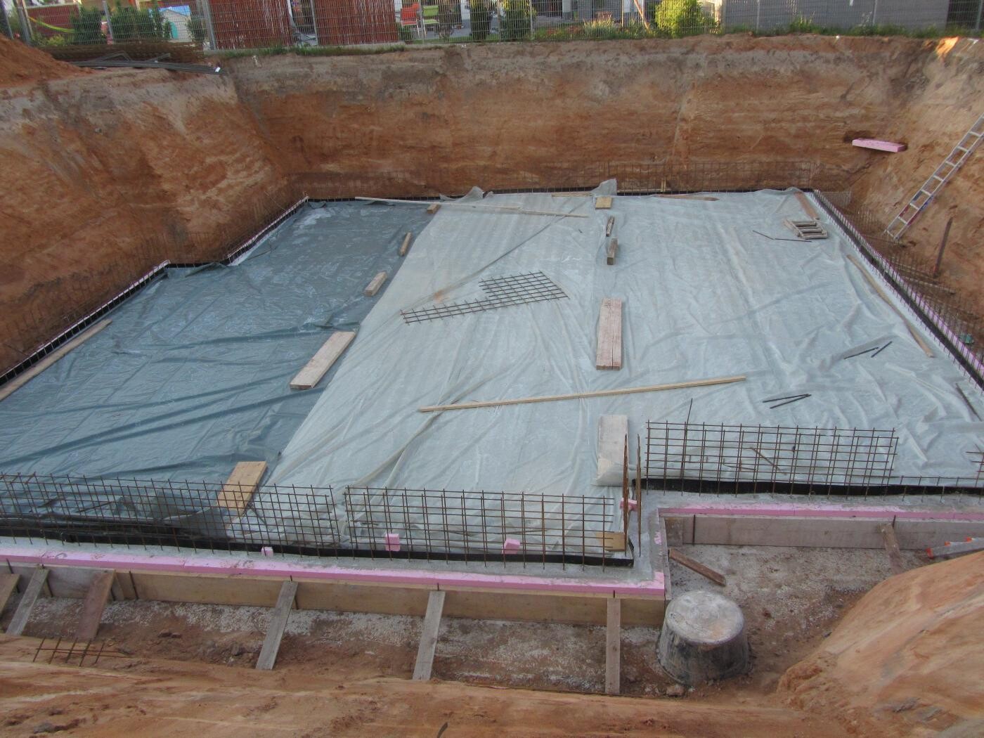 Fundamentgrube mit Abdeckfolie, Bewehrungsstahl, Holzlatten und Bauwerkzeugen auf der Baustelle.