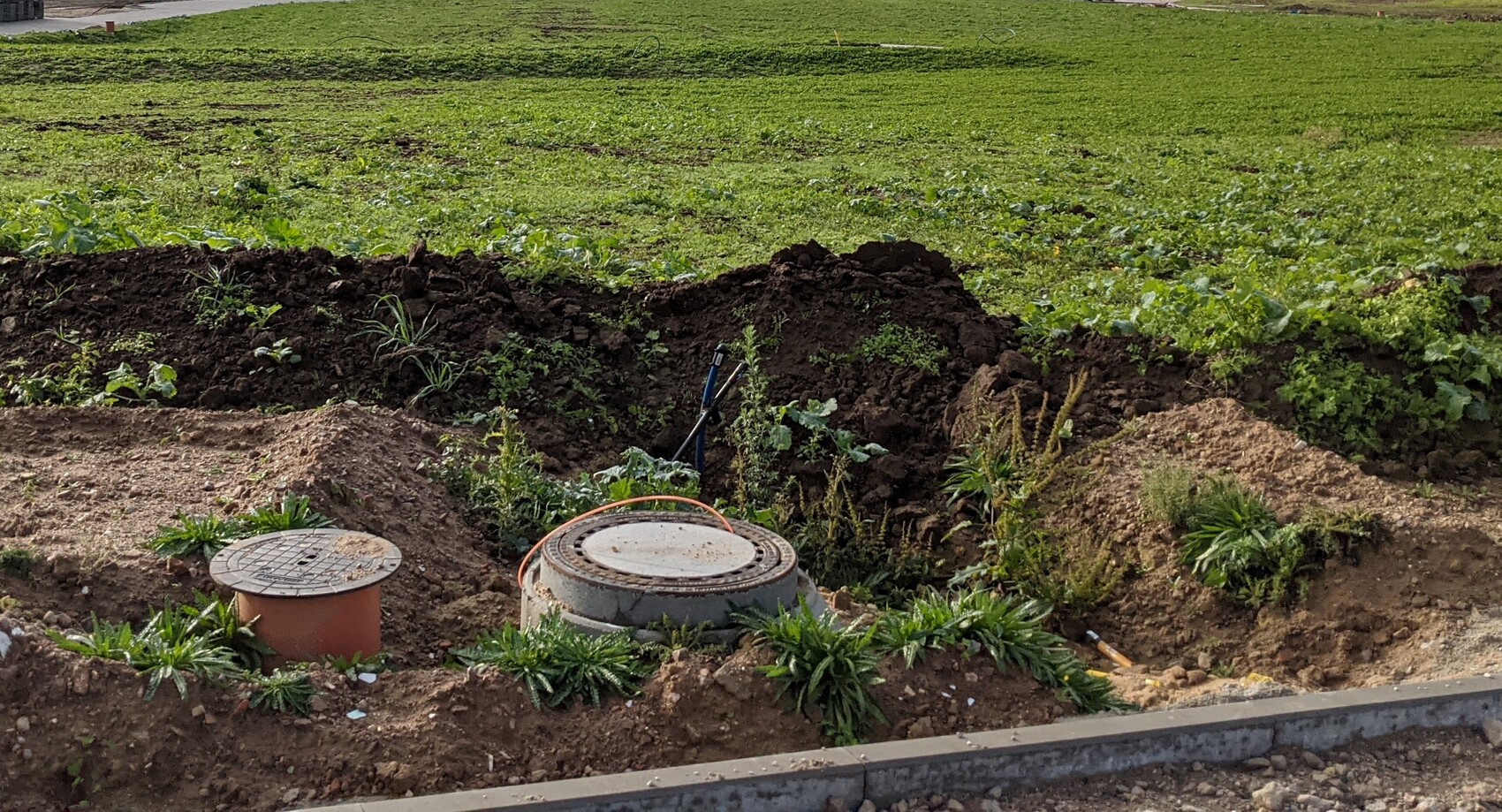 Offene Baugrube mit rundem Kanaldeckel, Erdwall und grünem Feld im Hintergrund.