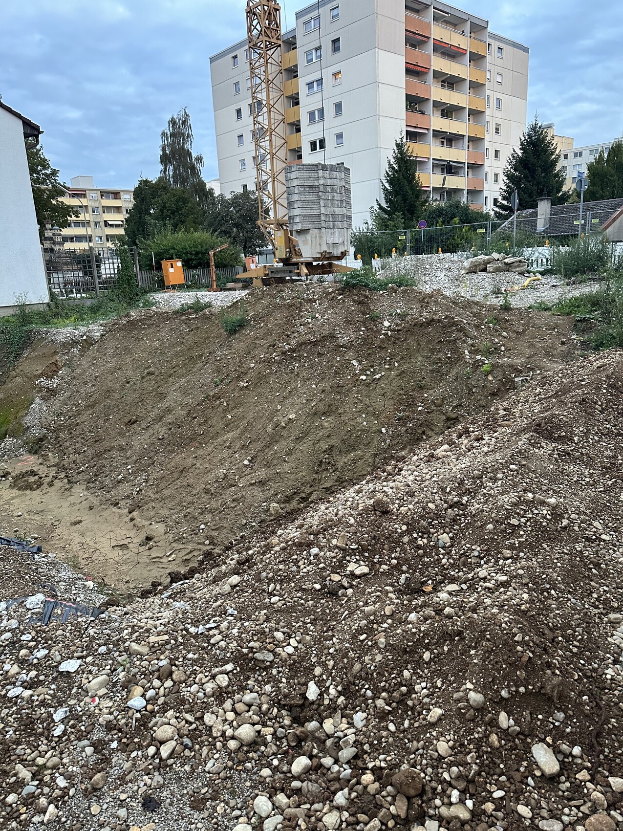Baustelle mit Erdhügel, Kran und Betonblöcken, Hochhaus im Hintergrund.