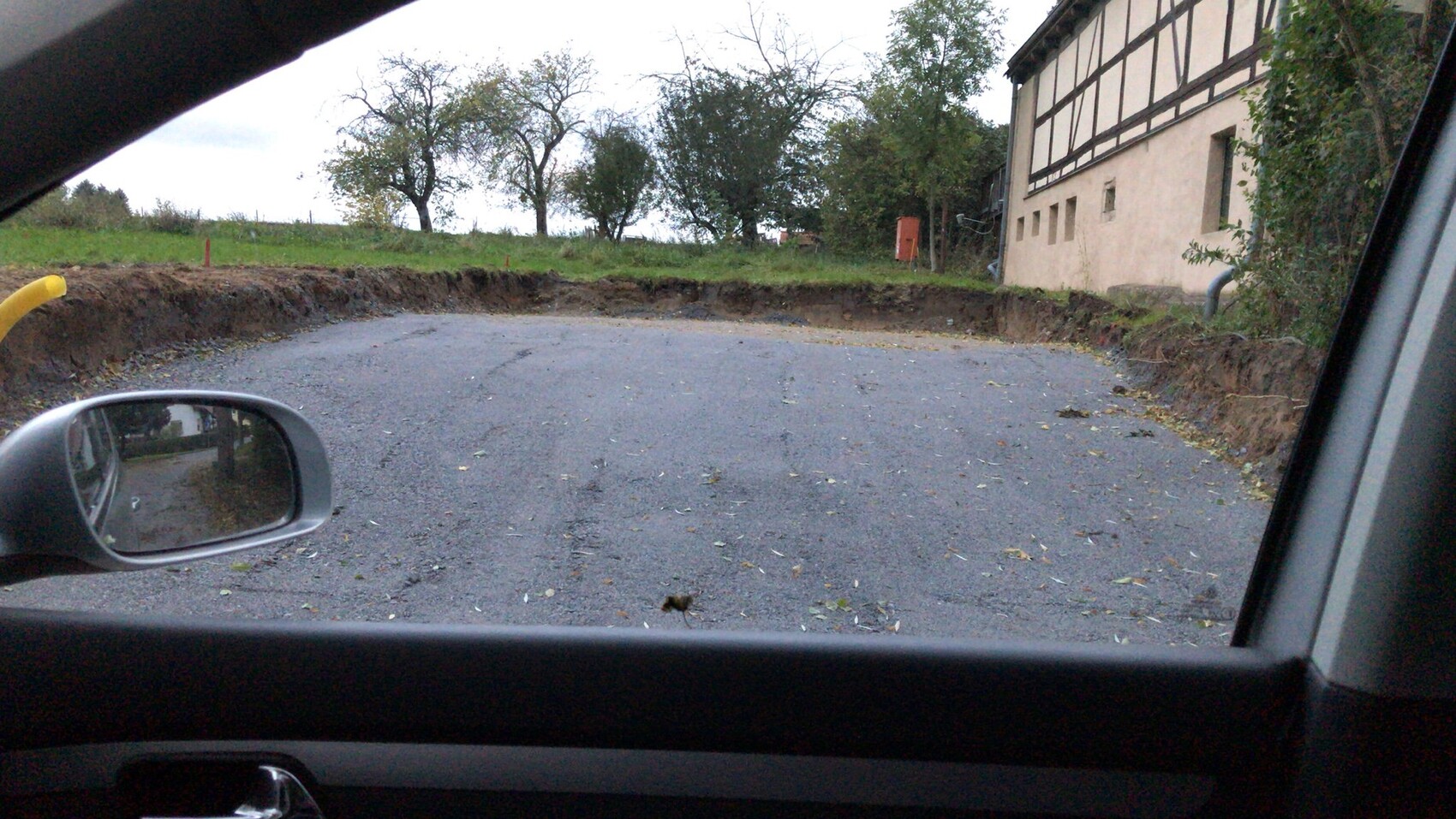 Blick aus dem Autofenster auf eine unbefestigte Kiesfläche neben einem Fachwerkhaus, Bäume dahinter.