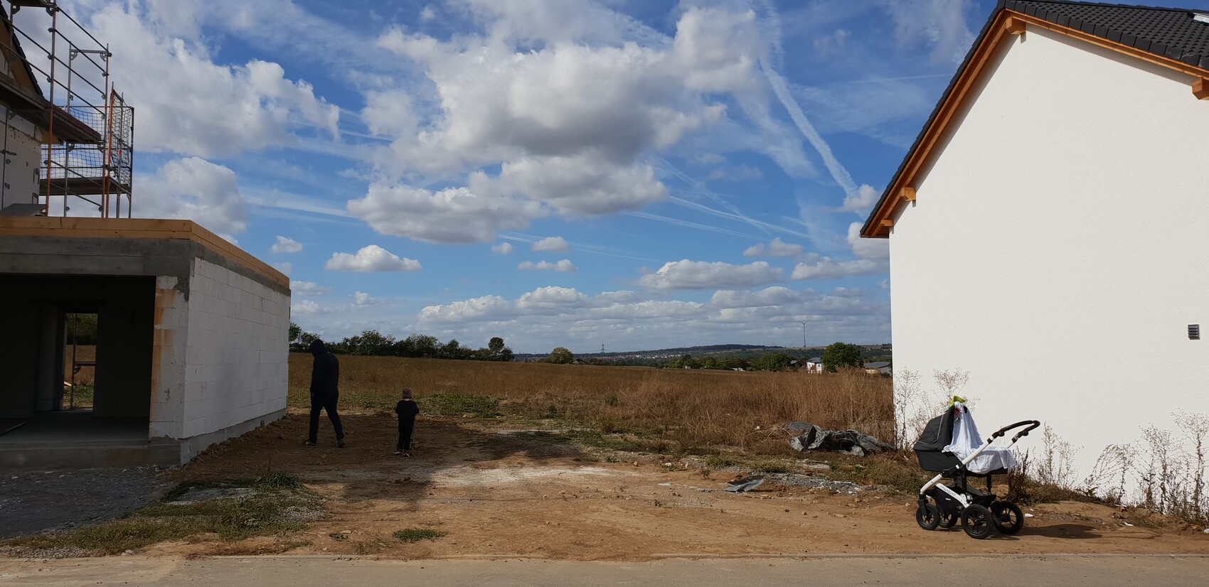 Baustelle links mit Gerüst; zwei Personen gehen, rechts ein weißes Haus, Kinderwagen im Vordergrund.