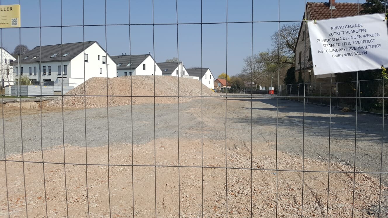 Baustelle hinter Drahtzaun mit Erdhaufen, Kiesfläche; Wohnhäuser im Hintergrund.