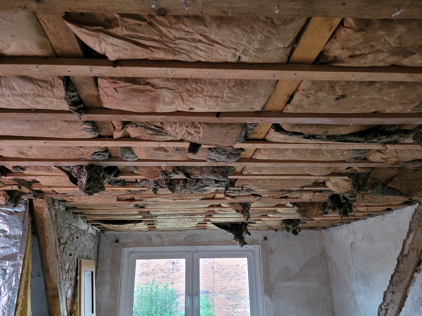 Zimmerdecke mit Holzbalken und loses Dämmmaterial; Fenster im Hintergrund