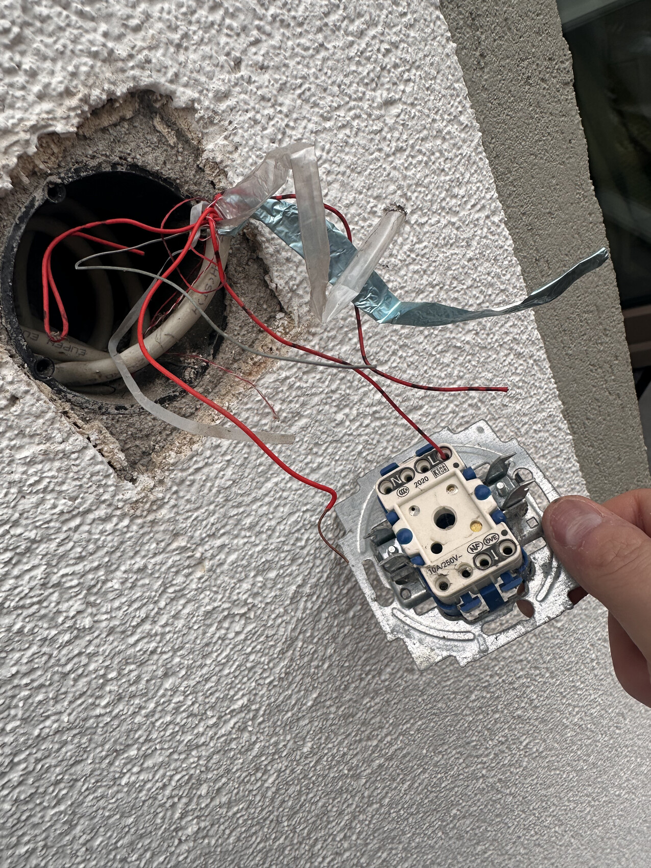Offene Wanddose mit roten und weißen Kabeln; Hand hält ein Steckdosen-/Schaltermodul.