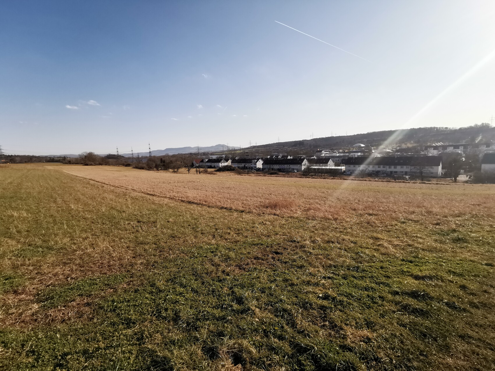 Großes Feld vor einem Dorf mit weißen Häusern am Hang; blauer Himmel und Kondensstreifen.