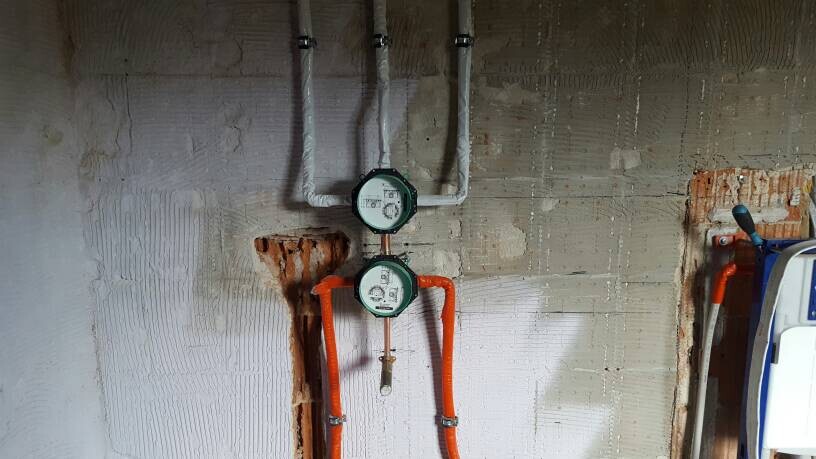 Zwei runde Wasserzähler an einer Wand, isolierte Rohre in Weiß und Orange, Heizungsraum.
