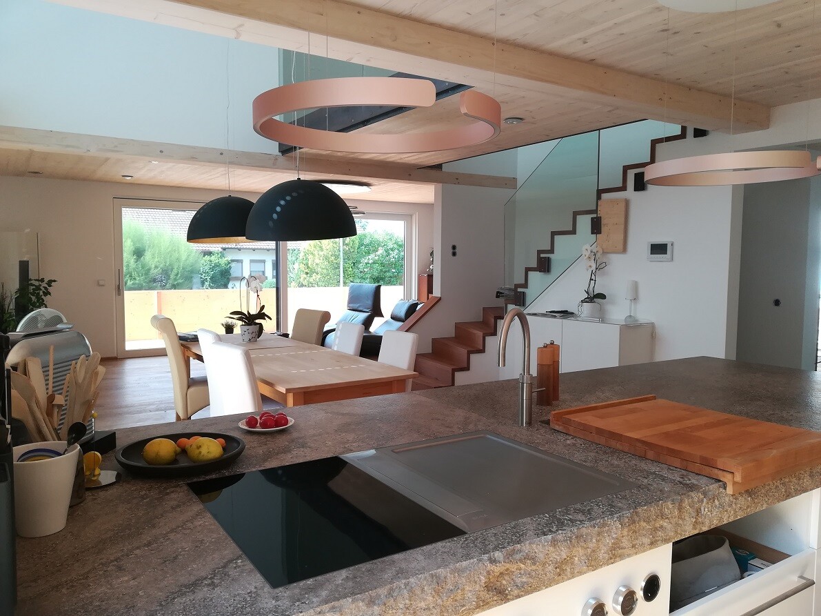 Offene Wohnküche mit Insel, Esstisch, Treppe, Holzdeckenbalken und runden Hängelampen.