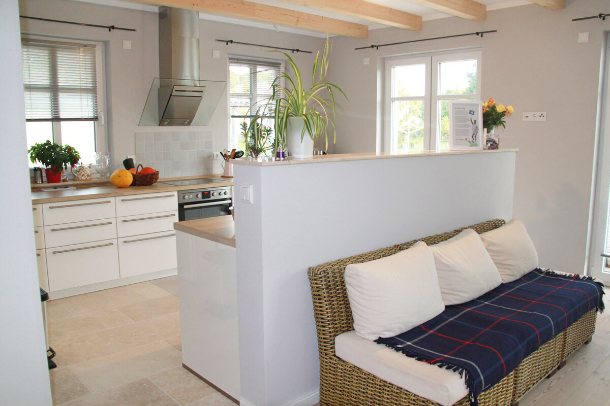 Offene Küche mit Insel, Dunstabzug und weißen Schränken; Korbsofa mit weißen Kissen im Wohnzimmer.