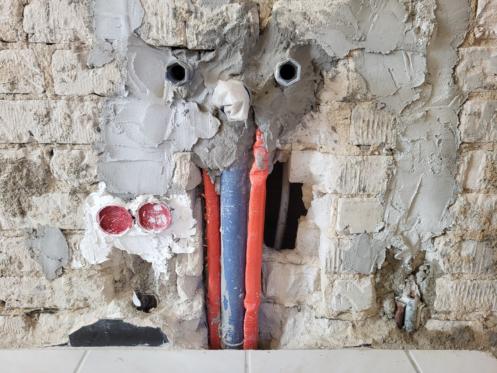 Offene rote und blaue Rohrleitungen in einer beschädigten Putzwand mit Löchern und Ziegeln.