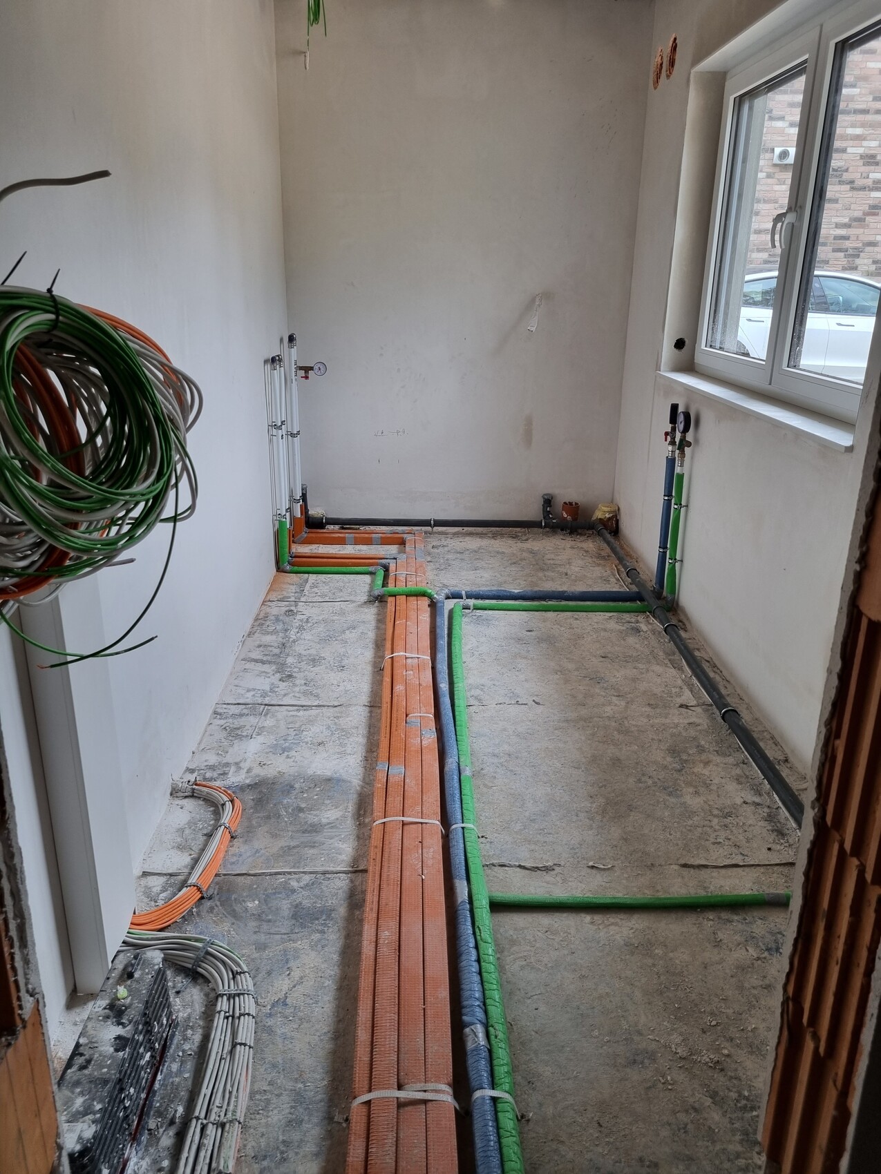Rohbau eines Zimmers mit verlegten farbigen Rohren (orange/grün/blau) auf dem Boden; Fenster rechts.