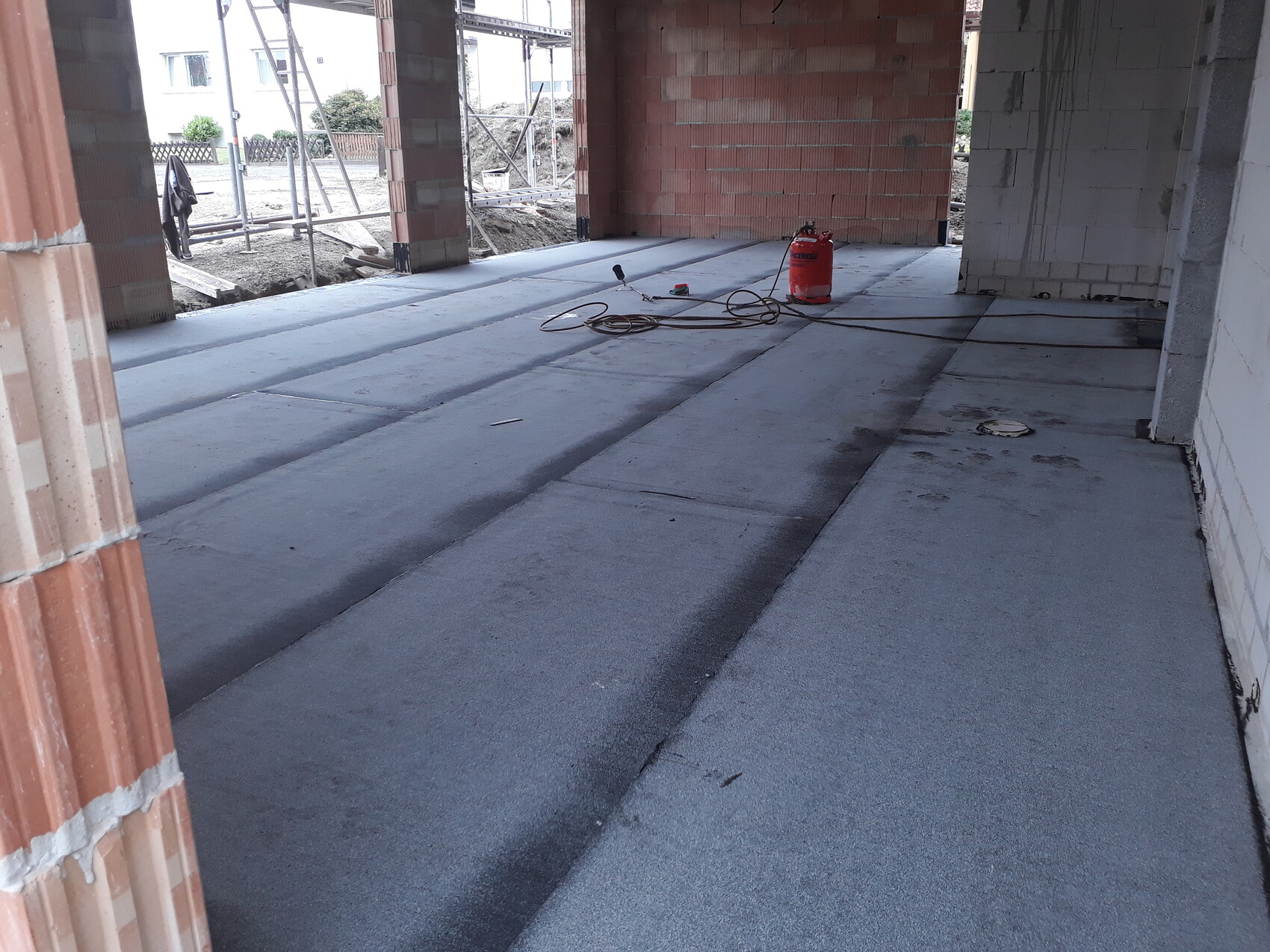 Innenraum-Baustelle: roter Feuerlöscher, Kabelsalat, Werkzeuge auf Betonboden, Backsteinwand.