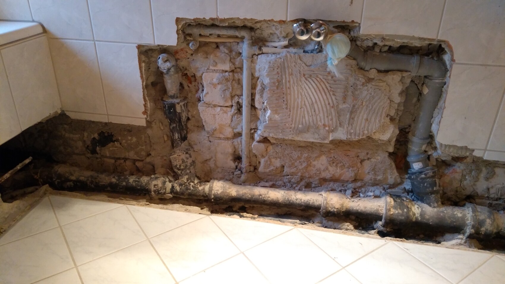 Offene Badezimmerwand mit freigelegten Rohren, Putzbruch und Fliesenresten.