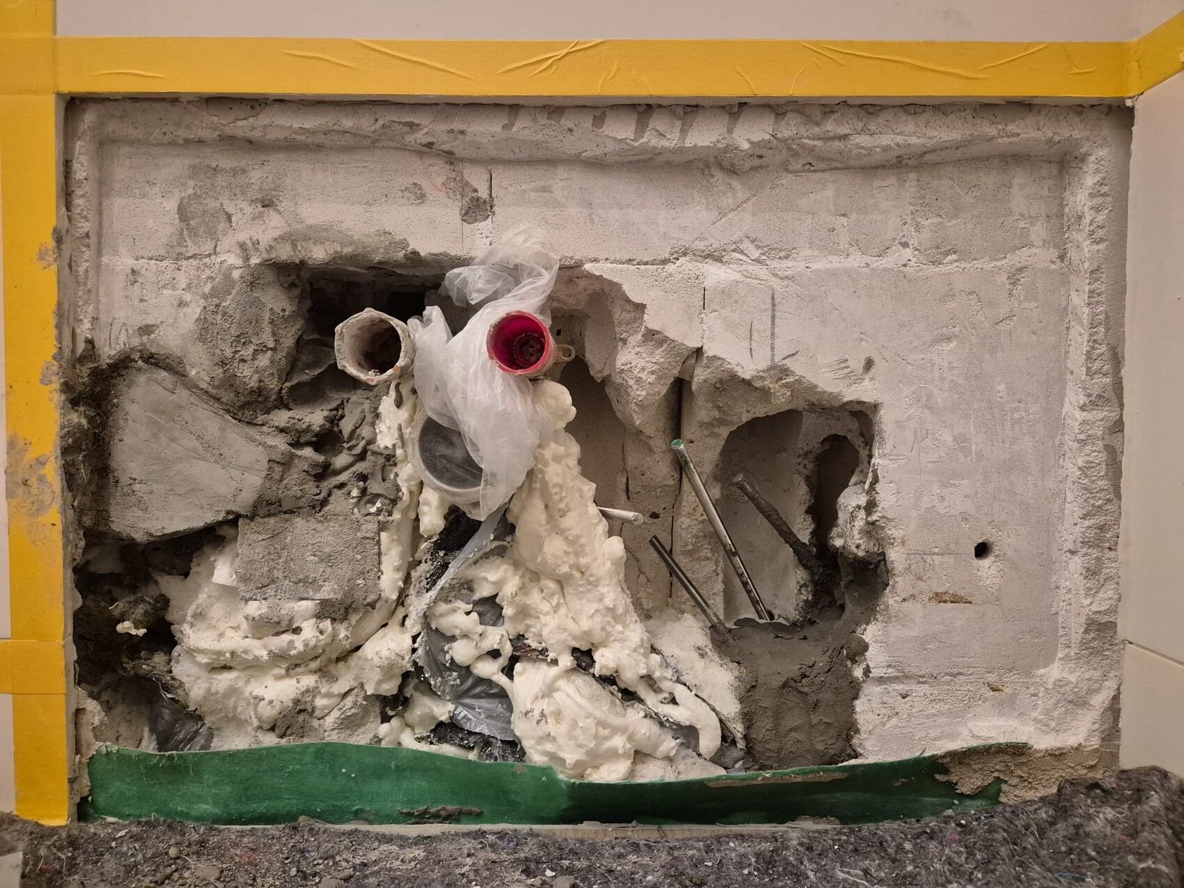 Freiliegende Sanitärrohre in Betonwand, Schaum-Daemmung und Bau-Reste; gelbe Rahmenmarkierung.