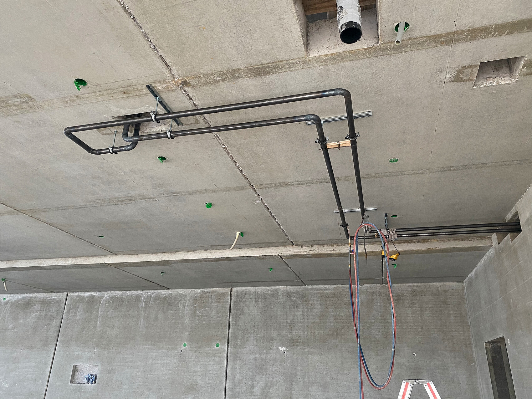 Schwarze Metallrohre an einer Beton-Decke einer Baustelle, blaue und rote Schläuche hängen herab.