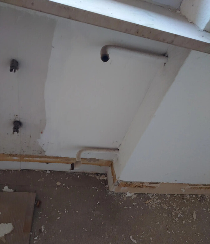 Weiße Kellerwand mit zwei Rohrleitungen, losem Putz und staubigem Boden.