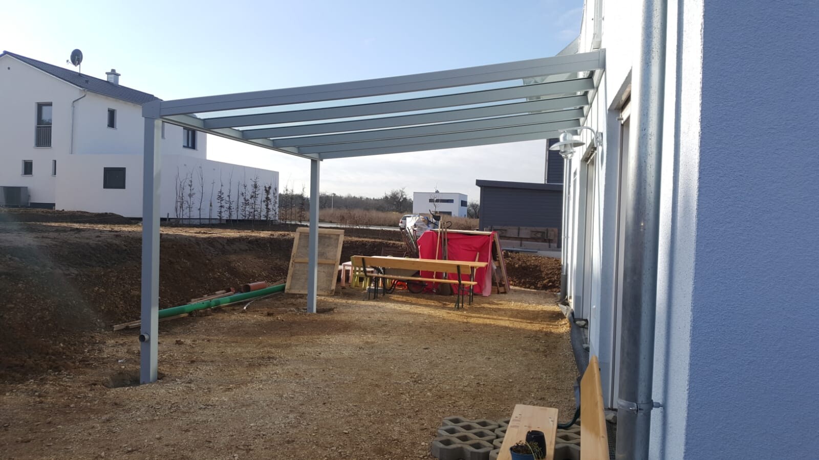 Offene Baufläche: Stahl-Pergola über Hof, Tisch und Bänke, Bauzubehör und Baustellenmaterial.