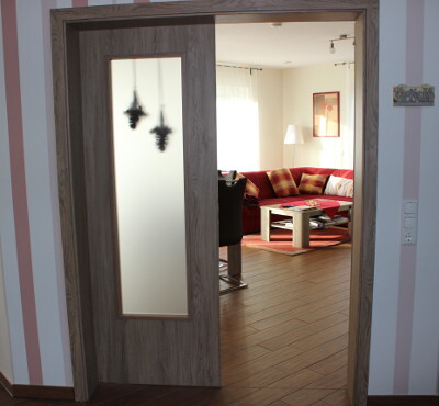 Offene Holztür mit Glasfeld führt zu modernem Wohnzimmer mit rotem Sofa.