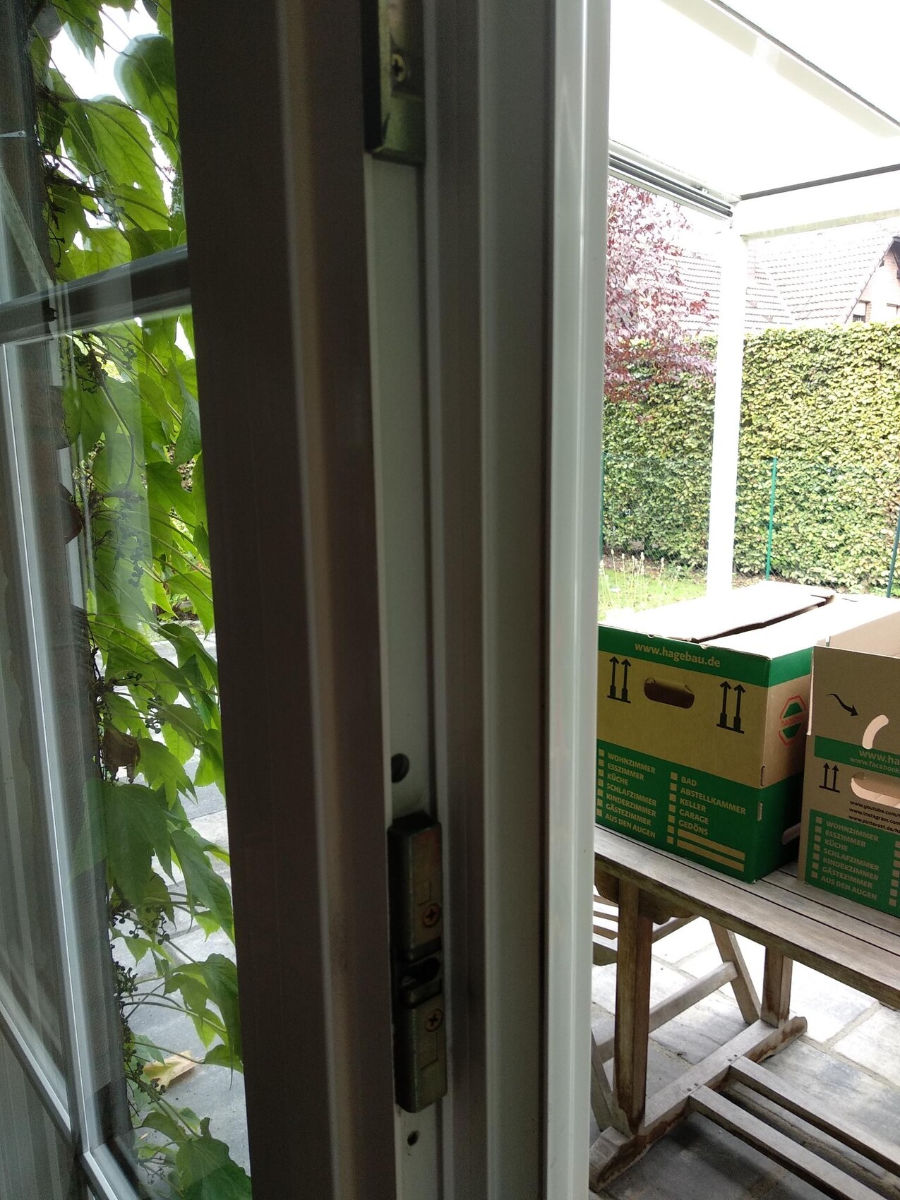 Offene Glas-Schiebetür; zwei Kartons stehen auf einer Holzbank draußen, grüne Hecke im Hintergrund.
