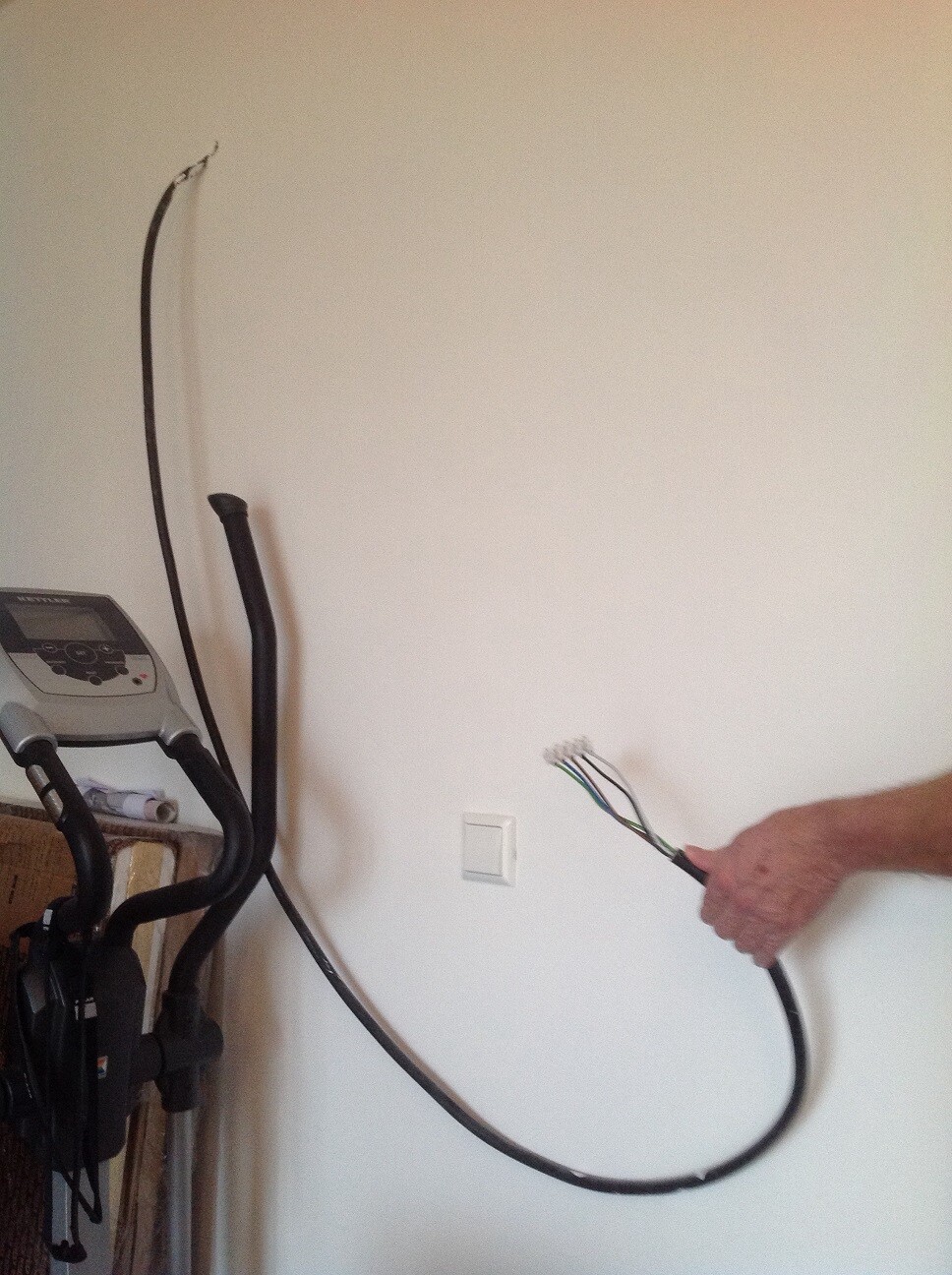 Hand hält lose Kabel mit mehreren Adern vor Wandsteckdose neben Heimtrainingsgerät