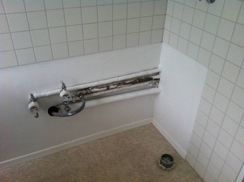 Badezimmer mit offenen Wandrohrleitungen, beschädigter Sanitärverkleidung und Fliesen.