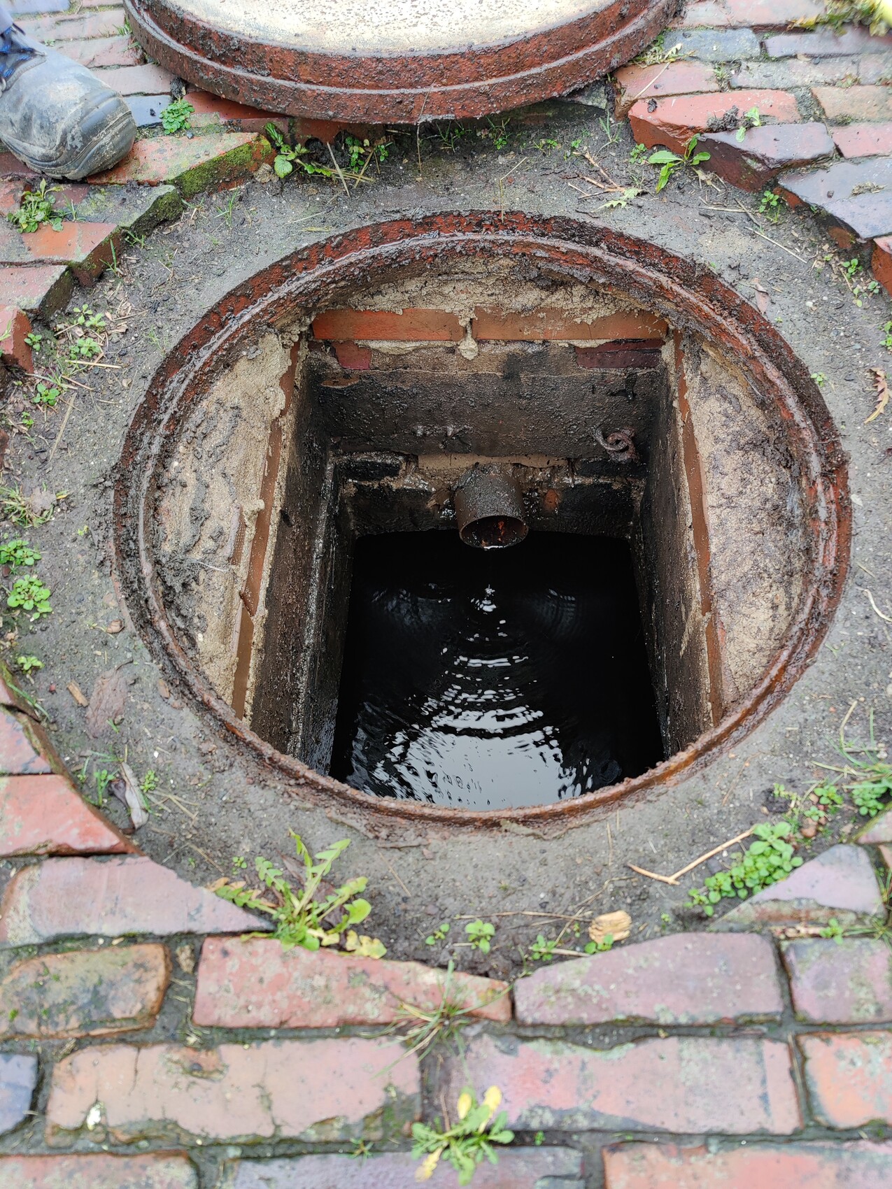 Offener Mannloch-Schacht mit dunklem Wasser, rostigem Rand, umgeben von Ziegel- und Pflastersteinen.