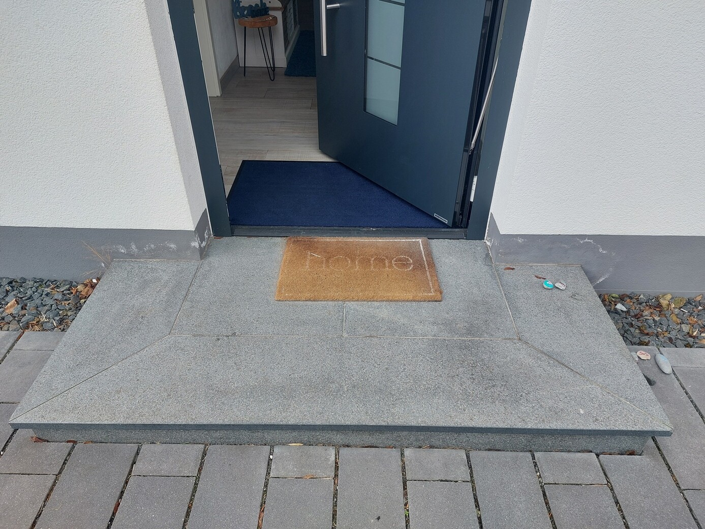 Offener Hauseingang mit blauer Tür, geöffneter Tür, grauen Steinplatten und Willkommensmatte.