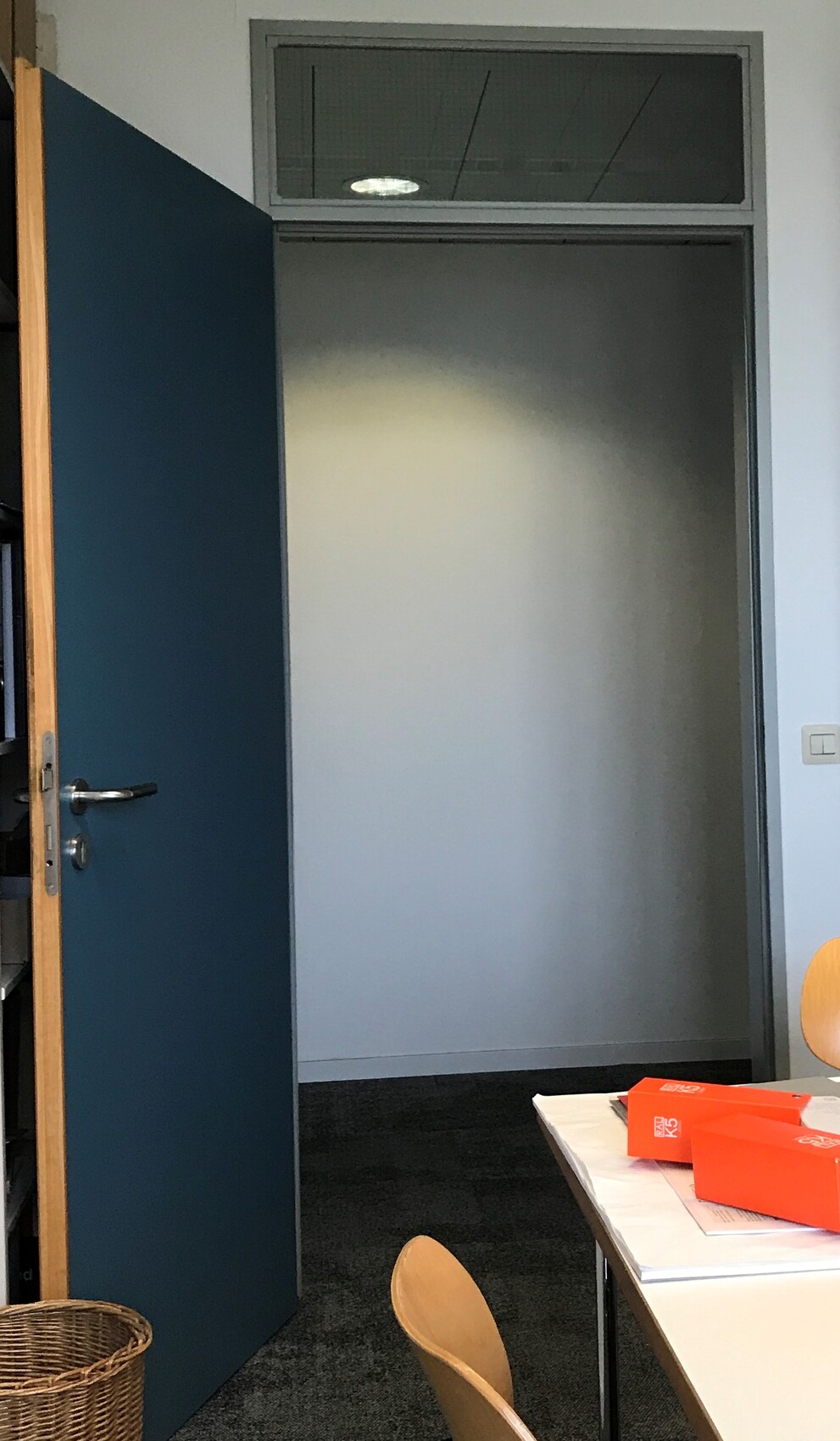 Offene blaue Bürotür; dahinter ein leerer Flur mit Oberlichtfenster über der Tür.
