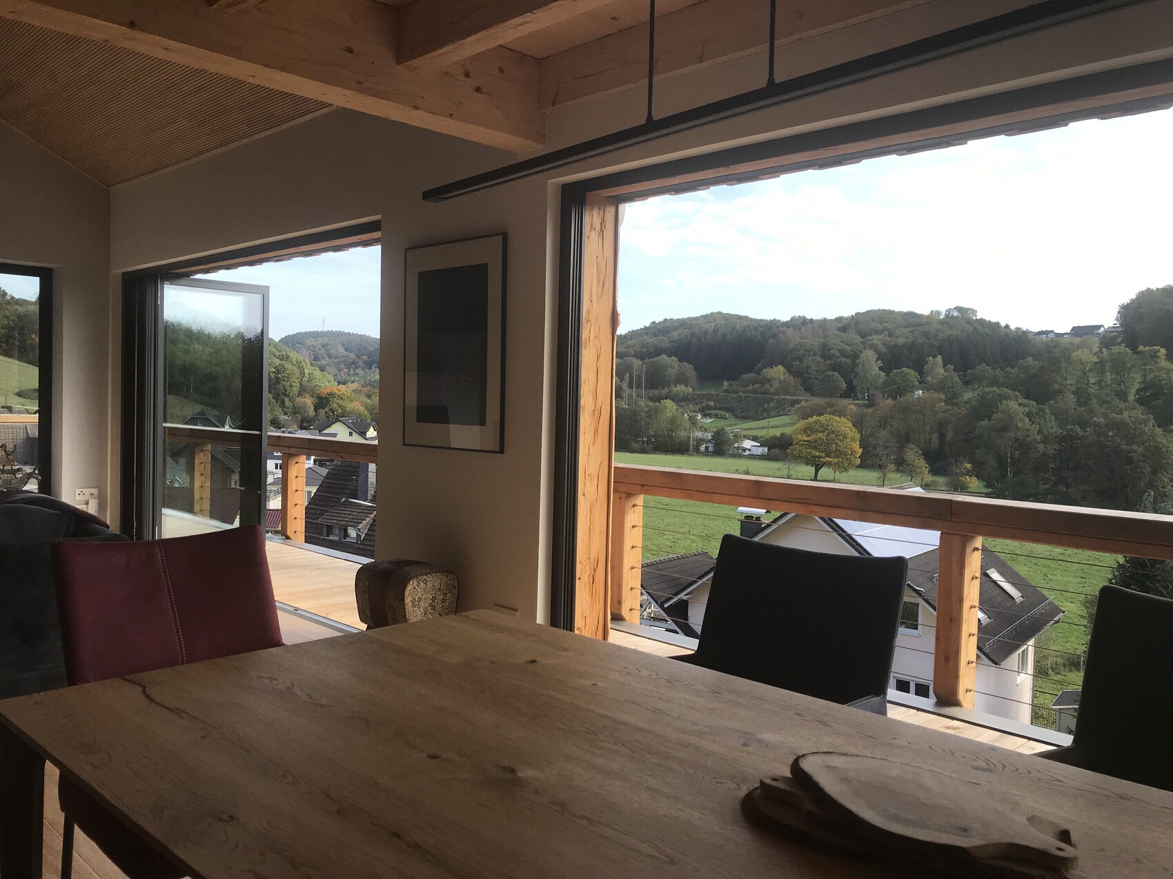 Wohnzimmer mit Holztisch, Glas-Schiebetüren und Balkonblick auf grüne Hügellandschaft.