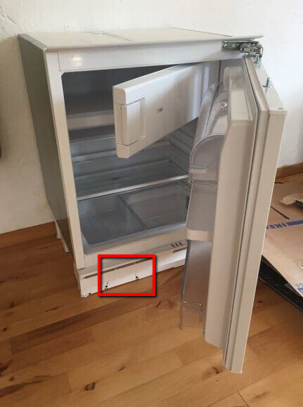 Weißer Kühlschrank mit offener Tür auf Holzboden; unten sichtbar lockeres Panel.