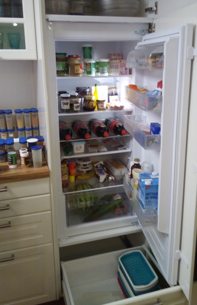 Offener Kühlschrank voller Gläser, Flaschen und Lebensmittel, Obst unten im Fach.