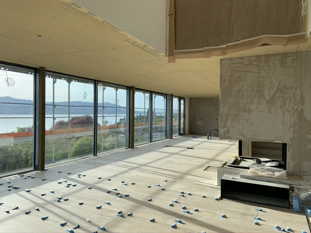 Große Glasfront mit Seeblick; unfertiger Innenraum, rohe Betondecke und Boden mit Fliesenkreuzen.