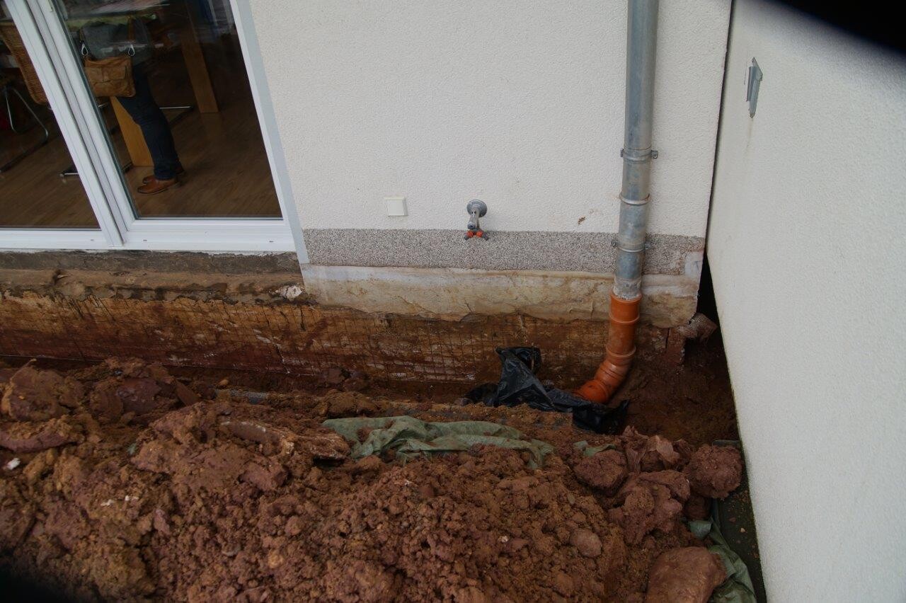 Außenansicht eines Hauses mit Baugrube, Rohrleitungen und Wasserhahn am Fundament.