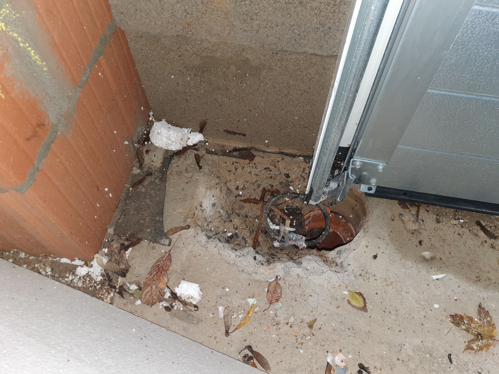 Offenes rundes Rohrloch im Boden neben dem Türrahmen, umgeben von Staub und Laub.