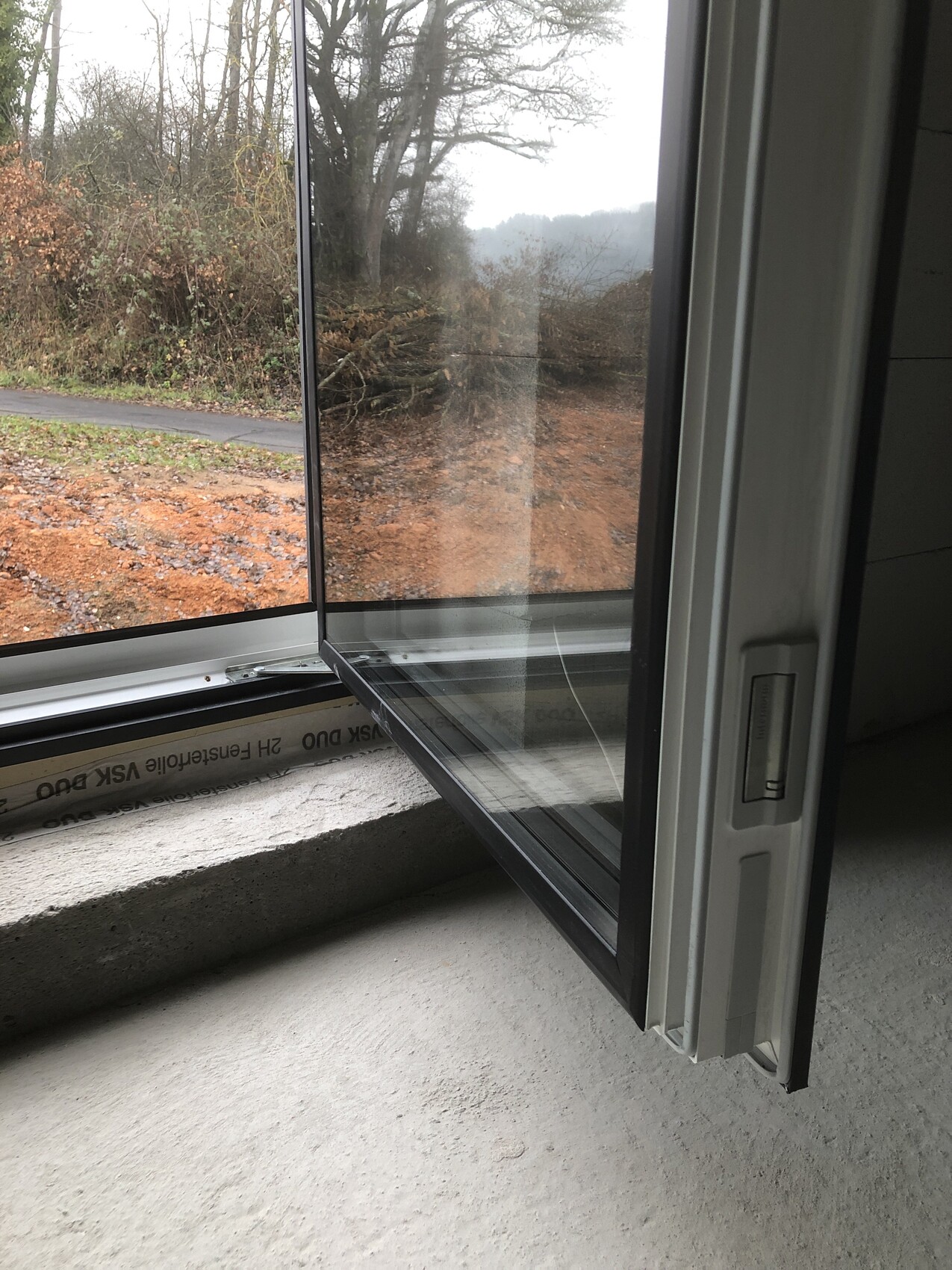 Offenes Fenster; Blick nach draußen auf Weg, Büsche und Bäume; Innenbetonboden erkennbar.